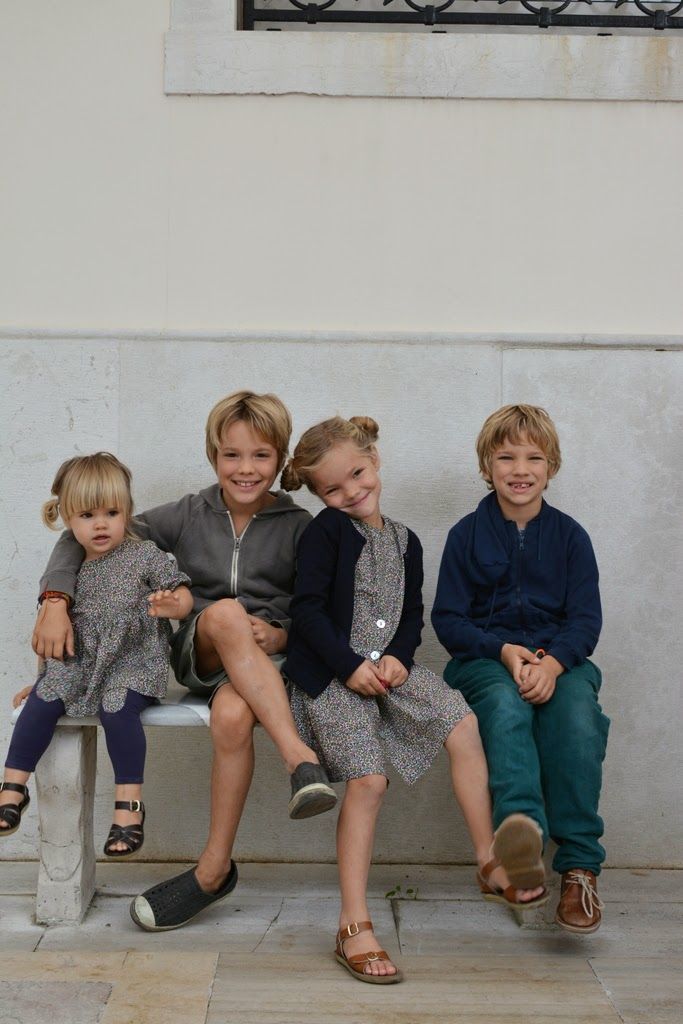 Four young children sitting on a bench, smiling and posing together against a light stone wall.