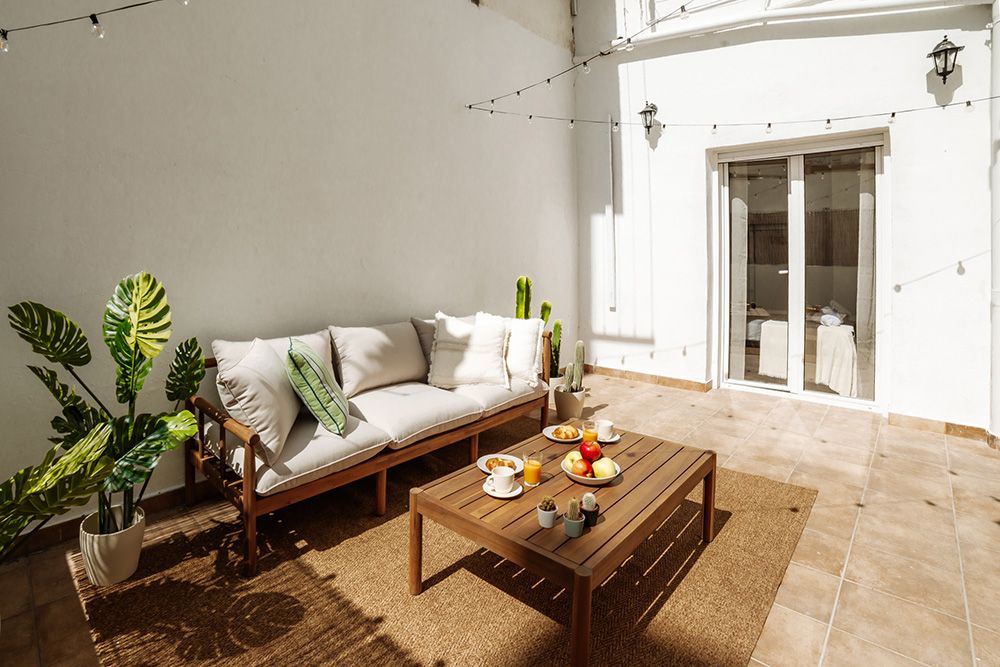 Sunny outdoor terrace with modern wooden sofa, coffee table set with breakfast, and potted plants against a white wall.
