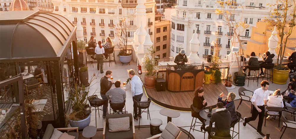 Rooftop bar in Valencia at sunset with people enjoying drinks, a DJ playing music, and views of historic buildings in the city center.