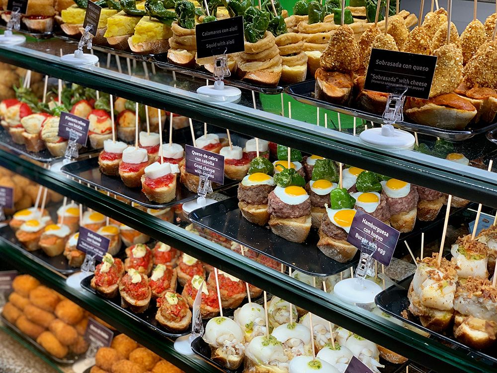 Display case filled with a variety of Spanish pintxos, including mini burgers with fried quail eggs, goat cheese with onions, squid with aioli, and other tapas served on bread with skewers.