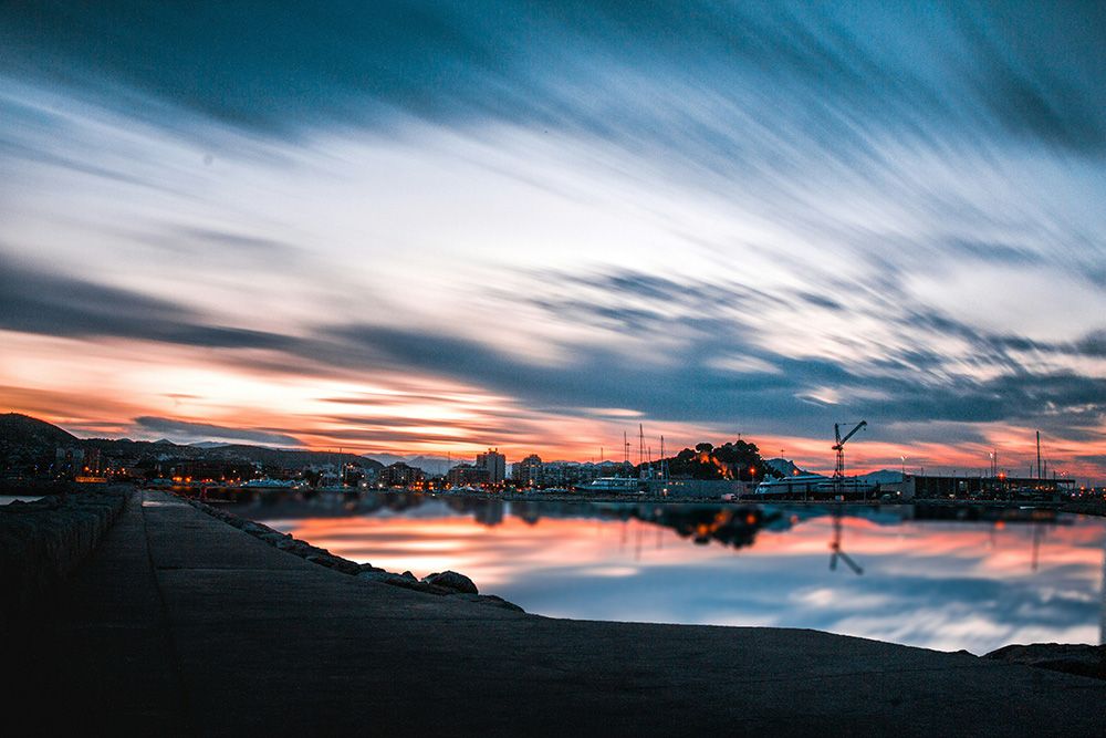 A serene coastal cityscape at sunset, with dramatic streaked clouds reflecting on calm water, silhouetted buildings and cranes along the shoreline, and a stone pier leading into the distance.