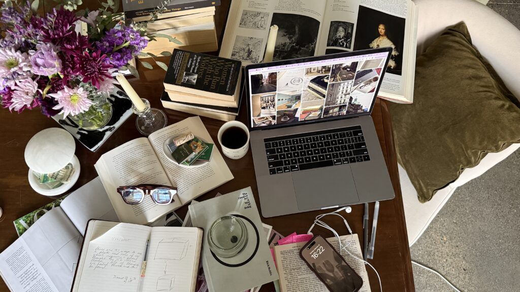 Top view of a cluttered desk with an open laptop, books, notebooks, glasses, coffee, and flowers, creating a busy workspace atmosphere