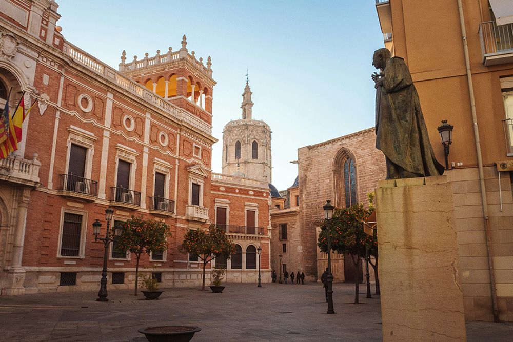 View of Plaza de la Virgen in Valencia with the bronze statue of a man in the foreground, the historic red-brick building of the Generalitat Palace on the left, and the bell tower of Valencia Cathedral rising in the background.