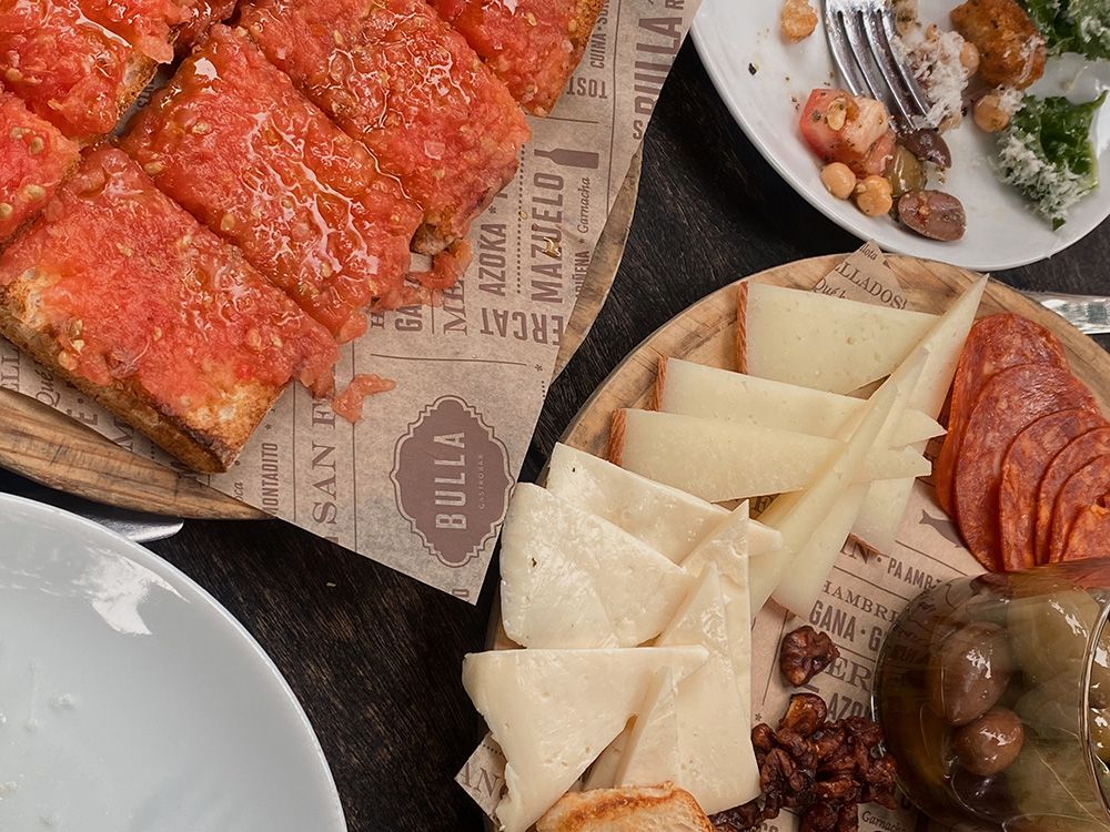 Spanish tapas spread with slices of toasted bread topped with tomato, a wooden board of manchego cheese and chorizo, olives in a glass jar, and a plate with salad remnants.