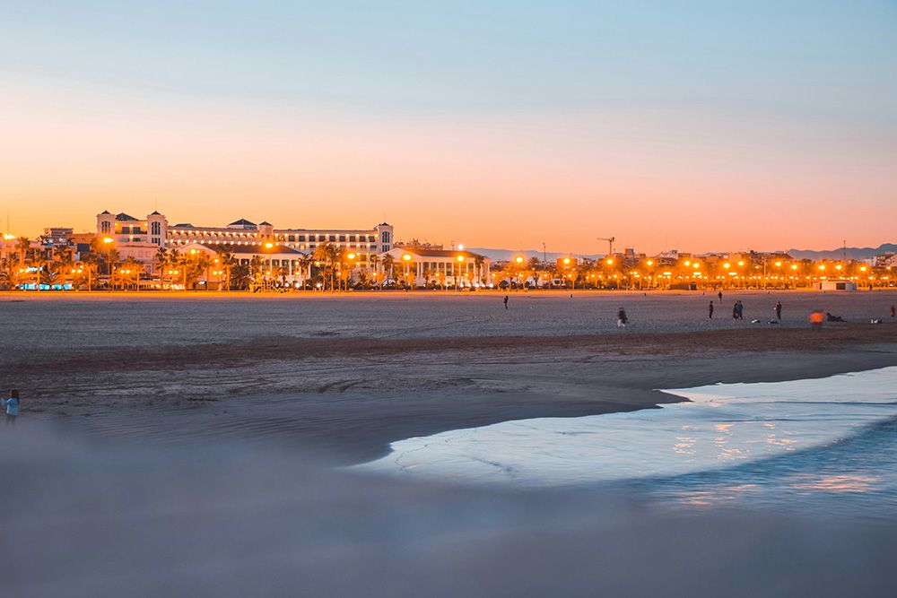 Evening view of Malvarrosa Beach in Valencia with the illuminated buildings and palm trees along the promenade, soft waves on the shore, and a colorful sunset sky.