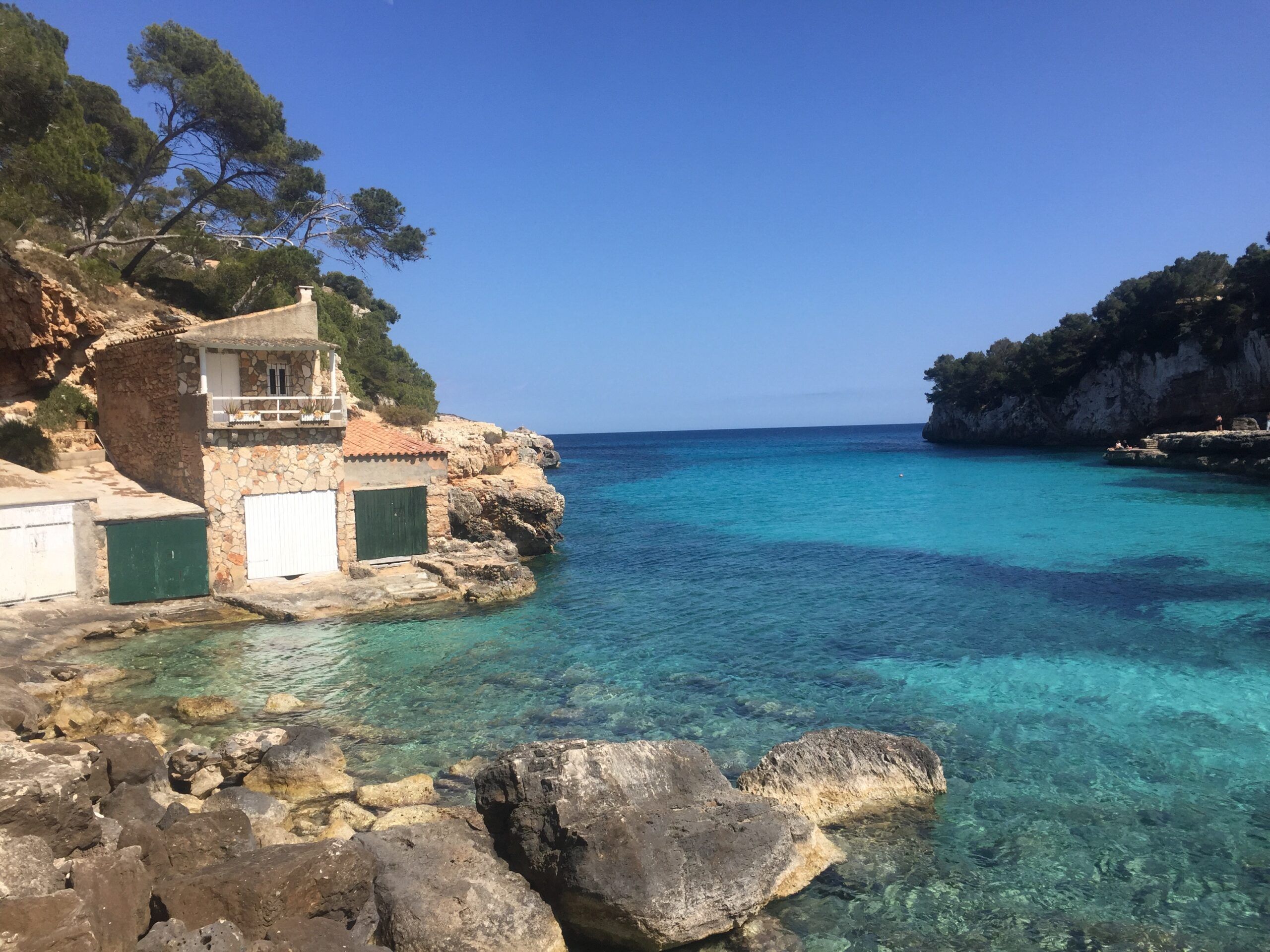Stone boathouses with green and white doors on a rocky shore overlooking a small turquoise bay surrounded by cliffs and pine trees under a clear blue sky.