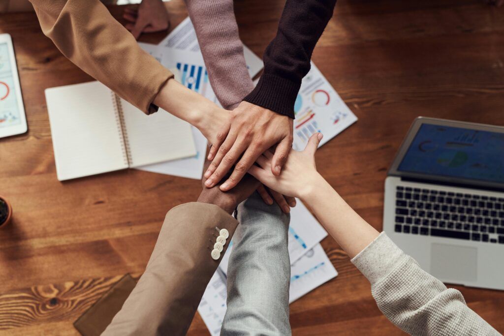 Group of diverse colleagues stacking hands together over a wooden desk with documents and a laptop, symbolizing teamwork and collaboration.