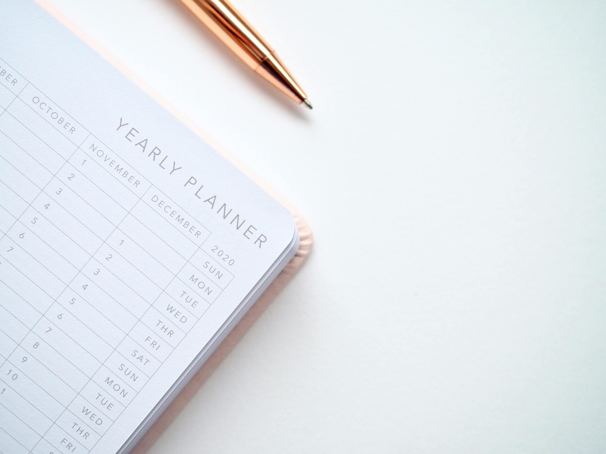 Close-up of a yearly planner open to the months of October to December with a gold pen placed beside it on a white desk.