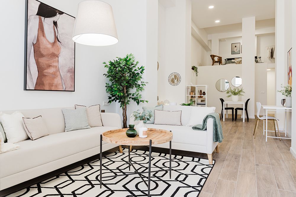 Bright modern living room with white sofas, a patterned rug, a round wooden coffee table, indoor plants, and wall art, leading into a dining area with high ceilings.