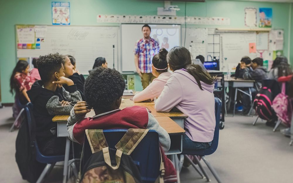 Students sitting at desks in a classroom listening to a teacher standing at the front near a whiteboard and projector screen.
