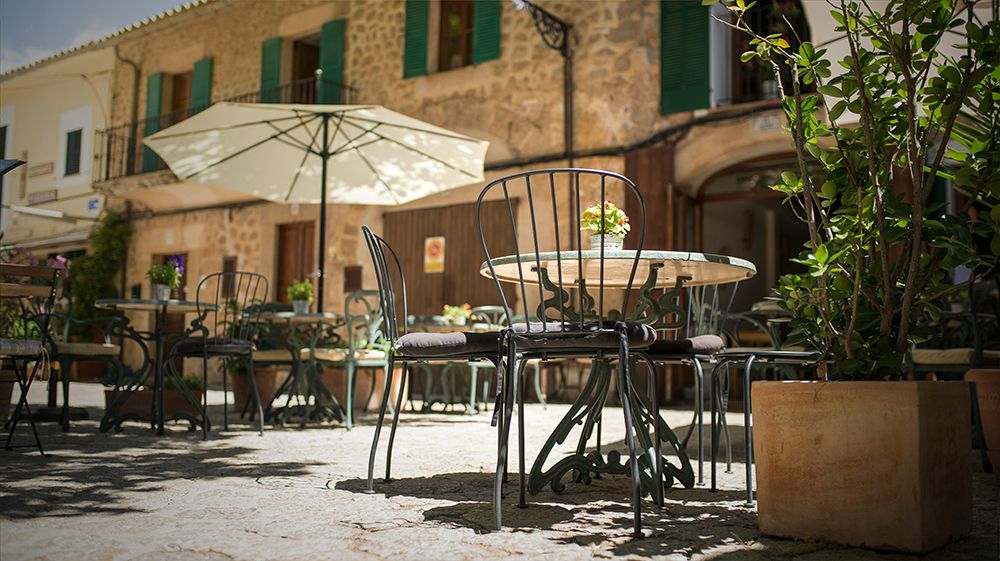 Outdoor café terrace in a charming Spanish village square with wrought-iron chairs, a round table, potted plants, and stone buildings in the background.