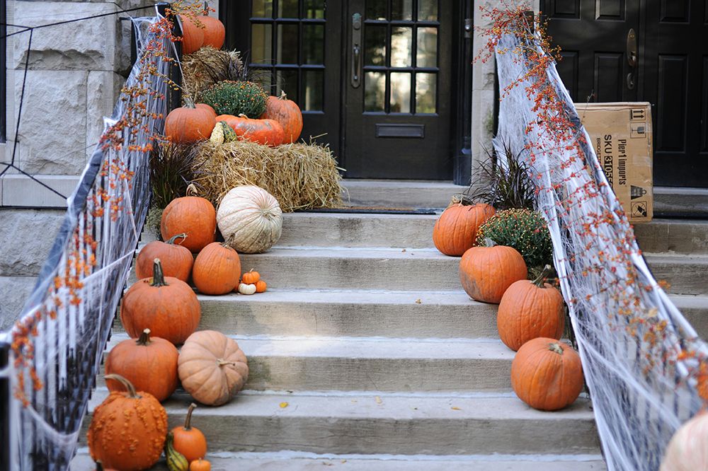Front steps decorated for Halloween with pumpkins, hay bales, cobwebs, and autumn plants leading up to a dark wooden door.