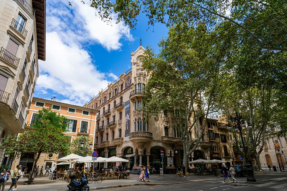 Street scene in Palma de Mallorca with people dining outdoors beneath umbrellas, framed by elegant historic architecture and trees.