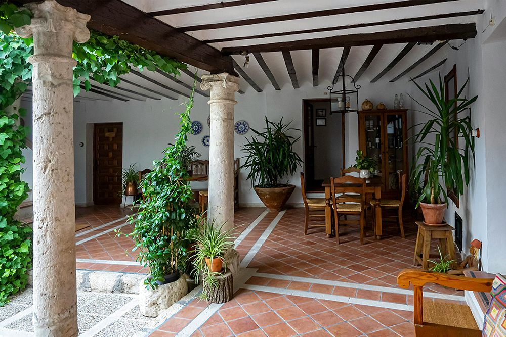 Traditional Spanish courtyard with terracotta tile flooring, stone columns, wooden beams, dining table, and lush green plants.