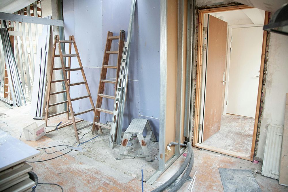Interior under renovation with ladders, construction materials, exposed pipes, and an unfinished wall.