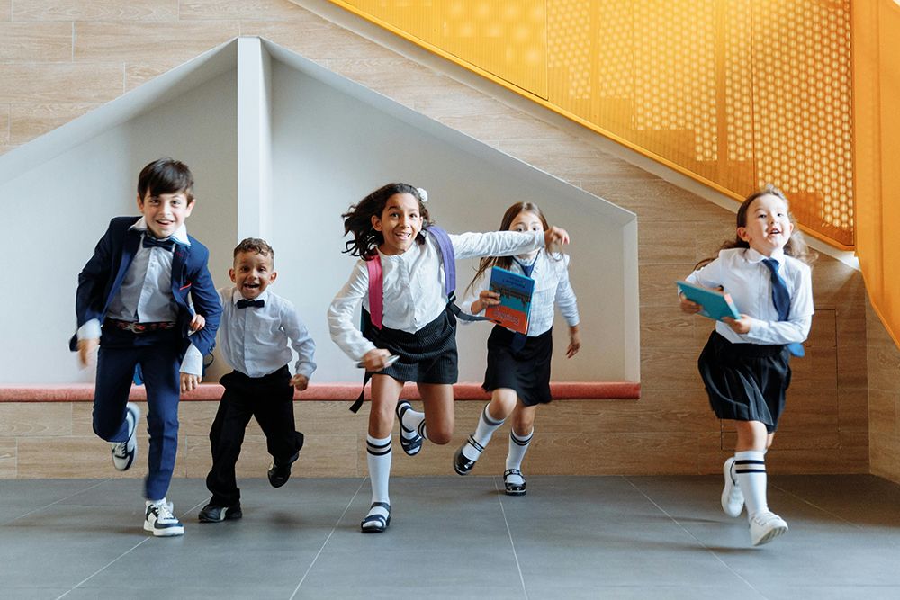 Group of young children in school uniforms joyfully running down a hallway, holding books and backpacks.