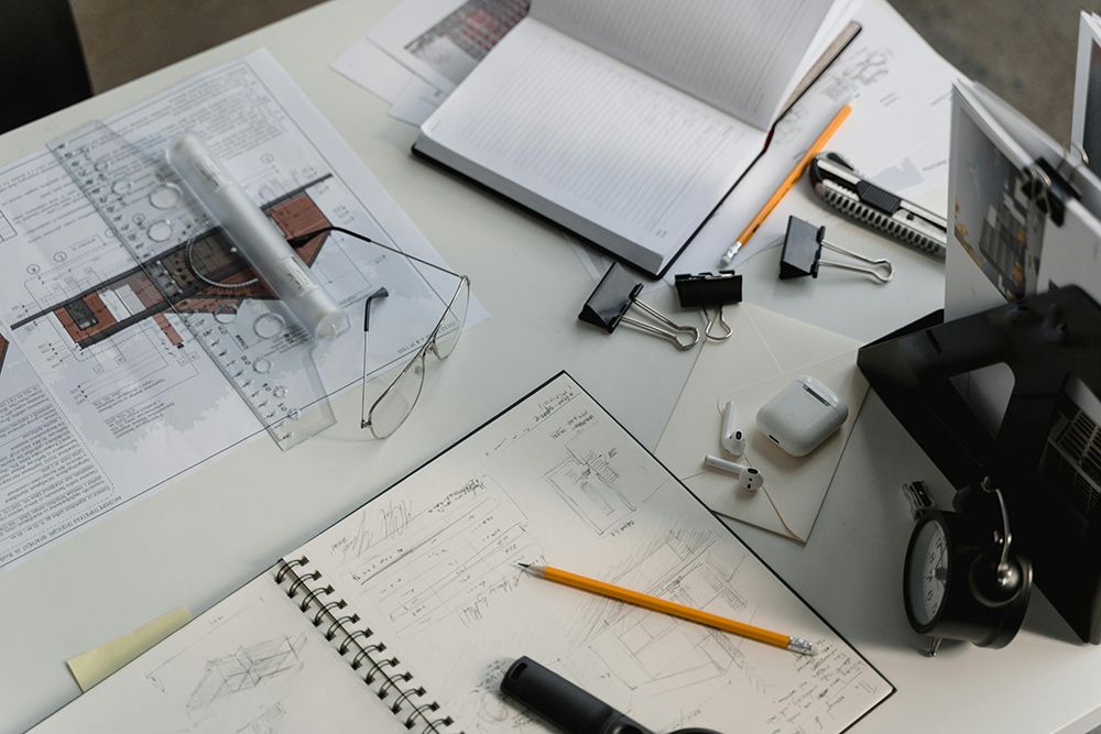 Cluttered workspace with architectural drawings, notebooks filled with sketches, drafting tools, glasses, and office supplies scattered across the desk.