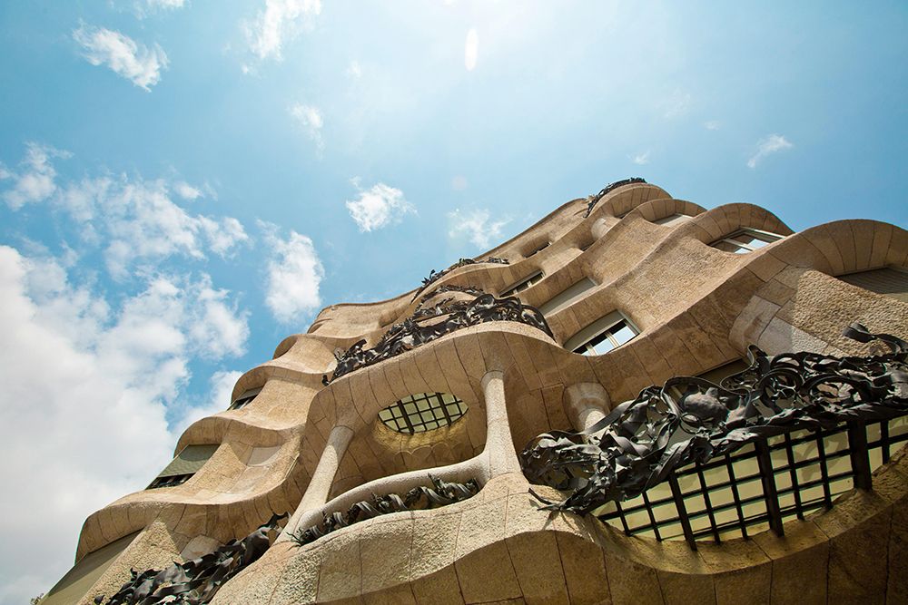 Upward view of Gaudí’s Casa Milà (La Pedrera) in Barcelona, showcasing its undulating stone façade and ornate iron balconies against a blue sky.