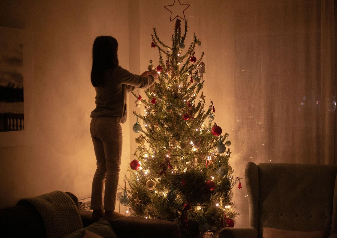 A young girl decorating a lit Christmas tree inside a cozy living room during the holiday season.