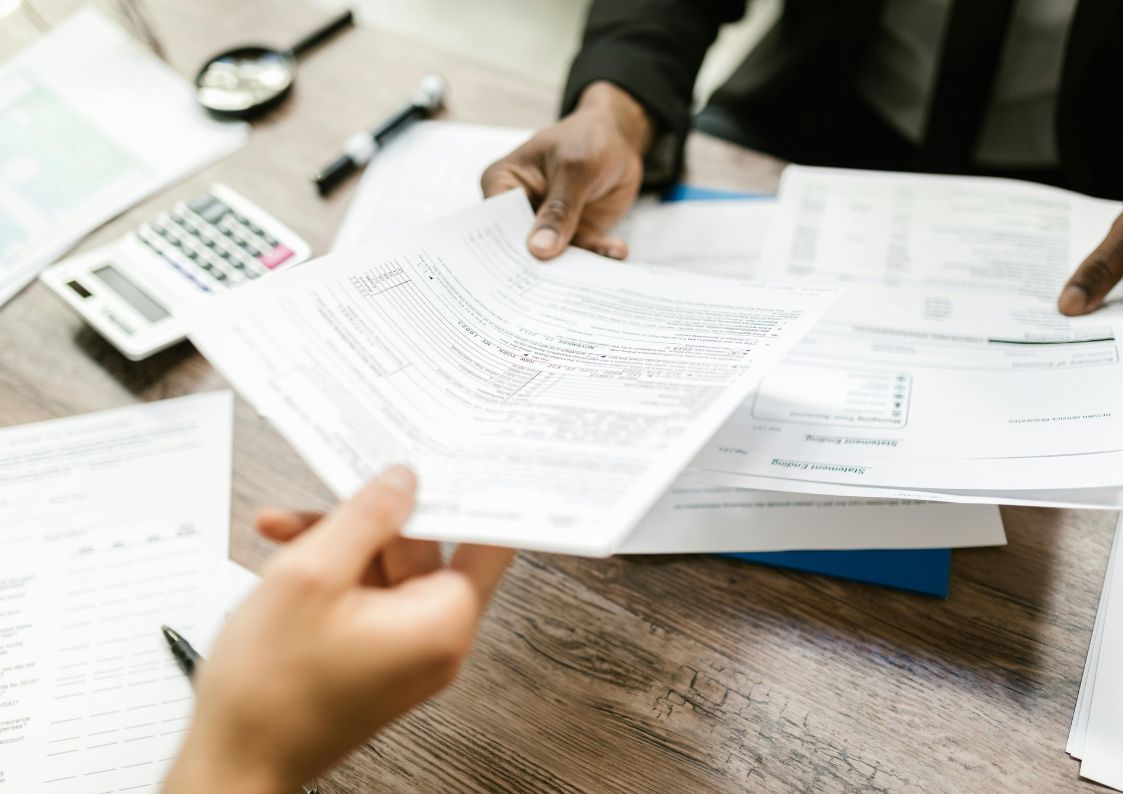 Two people exchanging and reviewing tax forms and financial documents at a wooden desk with a calculator nearby.