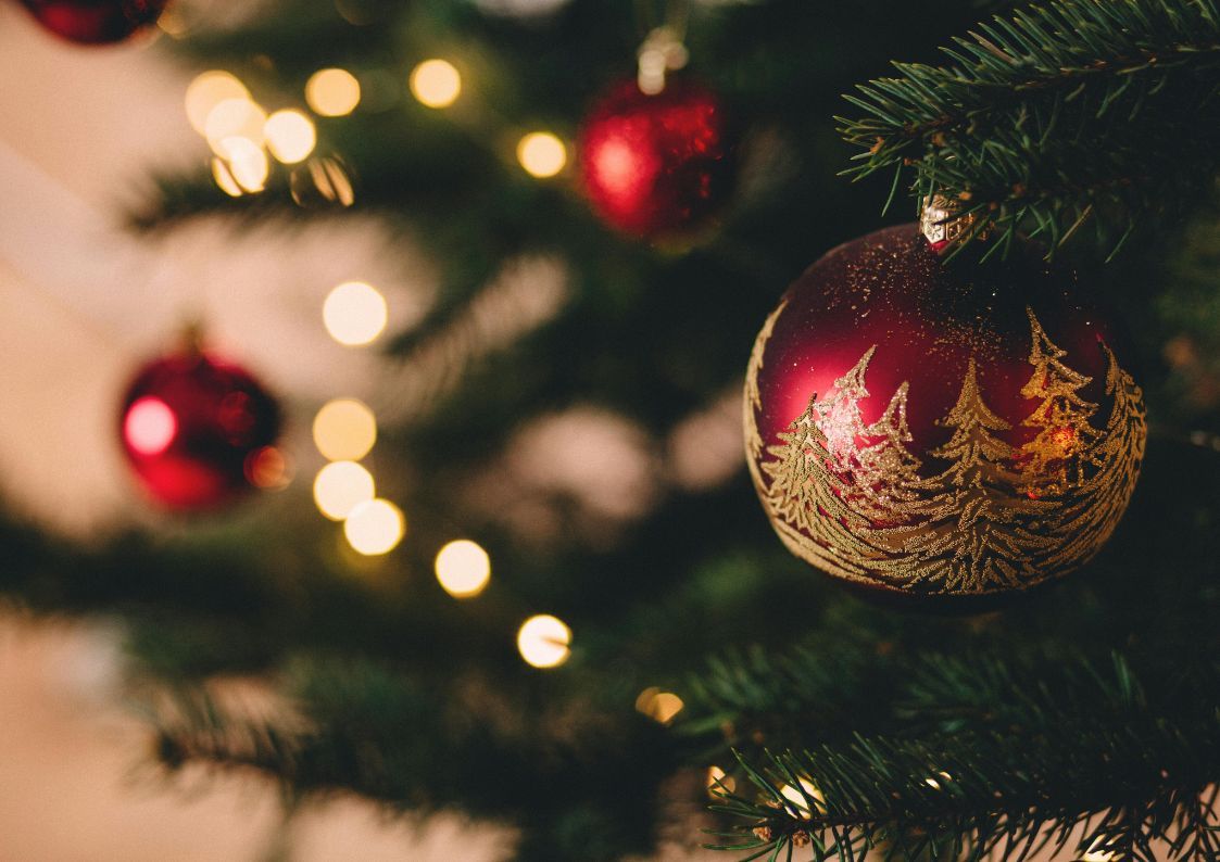 Close-up of a red Christmas bauble with gold tree designs hanging on a lit Christmas tree branch.
