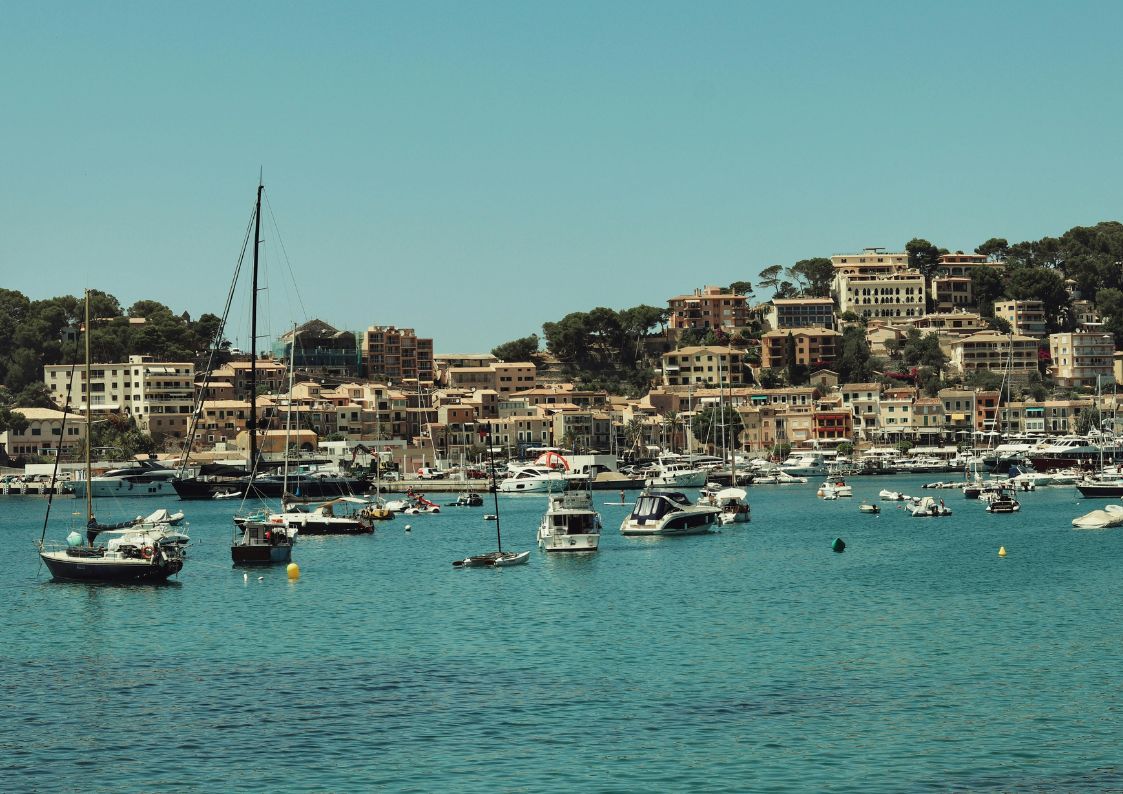 Coastal harbor town in Spain with boats moored in a marina and hillside homes overlooking the sea.
