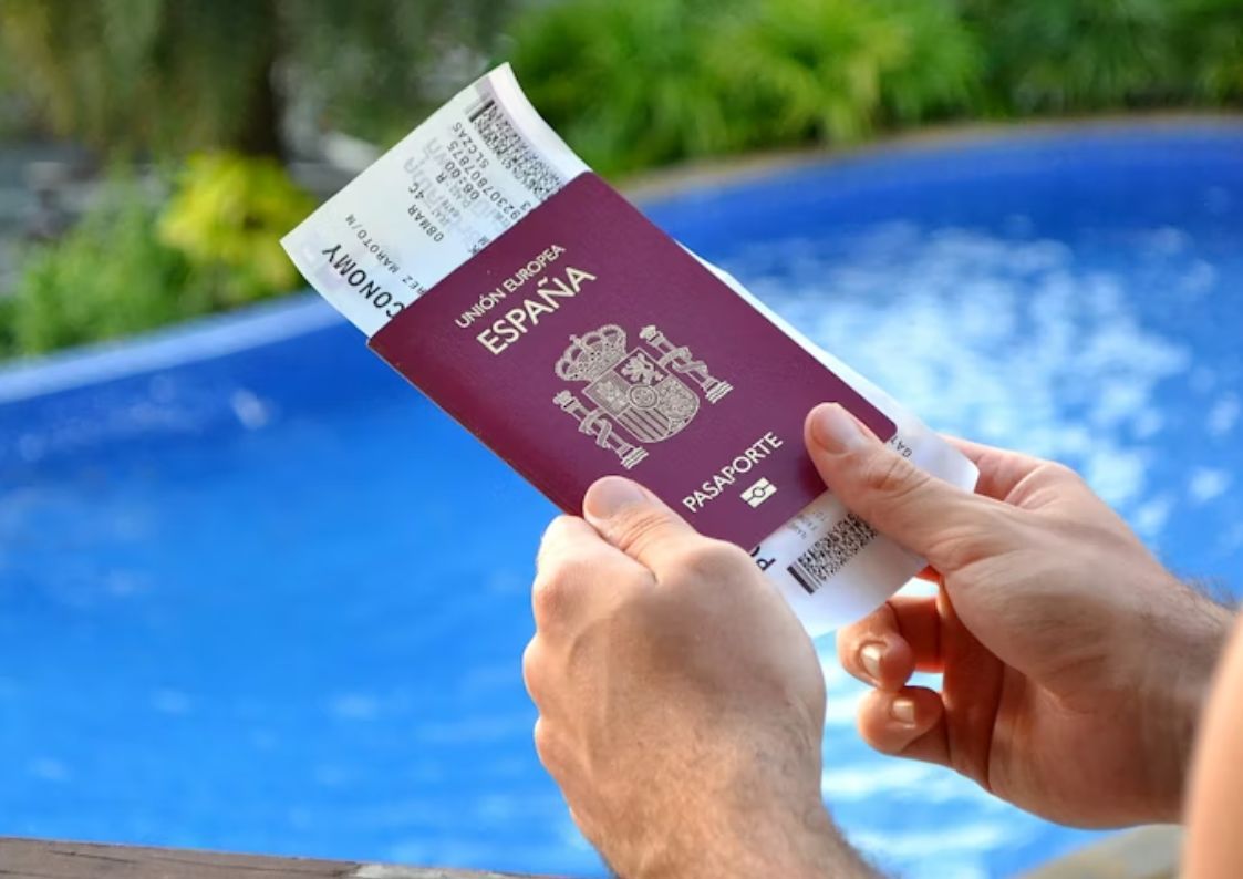 Hand holding a Spanish passport with boarding pass by a swimming pool, symbolizing residency, visas, and relocation to Spain.
